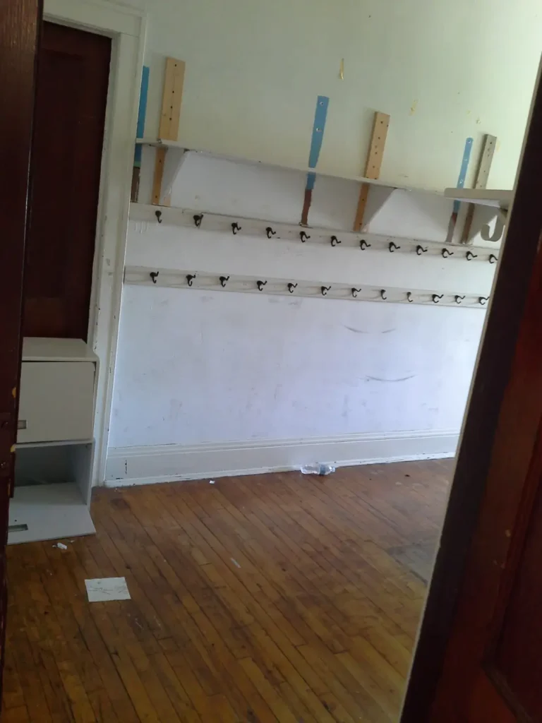 A narrow room for hanging coats and backpacks with peeling paint and scuff marks on the floor