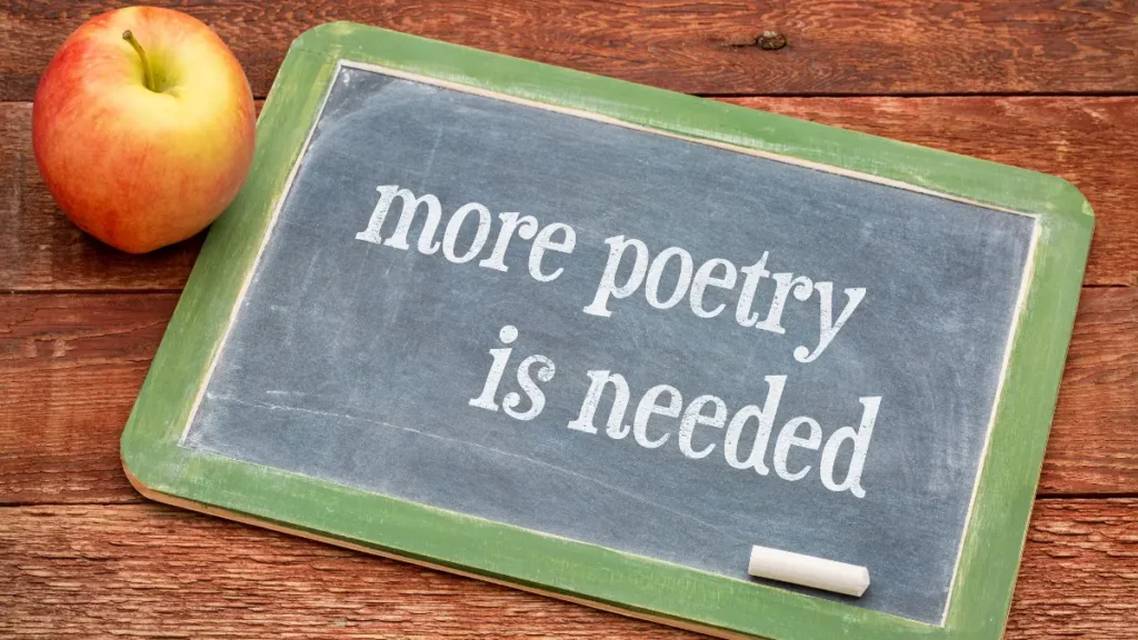 A chalkboard with the words "more poetry is needed" rests on a dark wood background beside a red apple and a piece of chalk